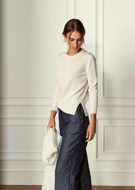 A woman wearing a cream long-sleeve knit sweater and dark blue wide-leg trousers, holding a matching cream bag against a white paneled wall.