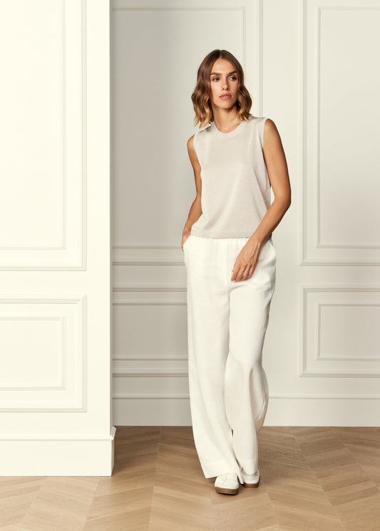 A woman stands in a bright interior with white paneled walls and light wood flooring, wearing a sleeveless light beige top and white wide-leg pants.