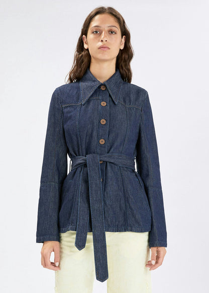 A woman modeling a dark blue denim belted jacket with brown buttons, paired with cream pants, against a plain light gray background.