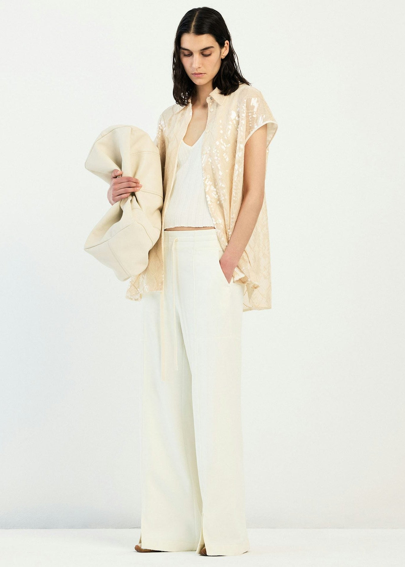 A model stands against a plain white studio background wearing a cream-colored outfit consisting of wide-leg trousers, a white top, a sleeveless cream vest with subtle gold sequins, and holding a matching cream draped wrap.