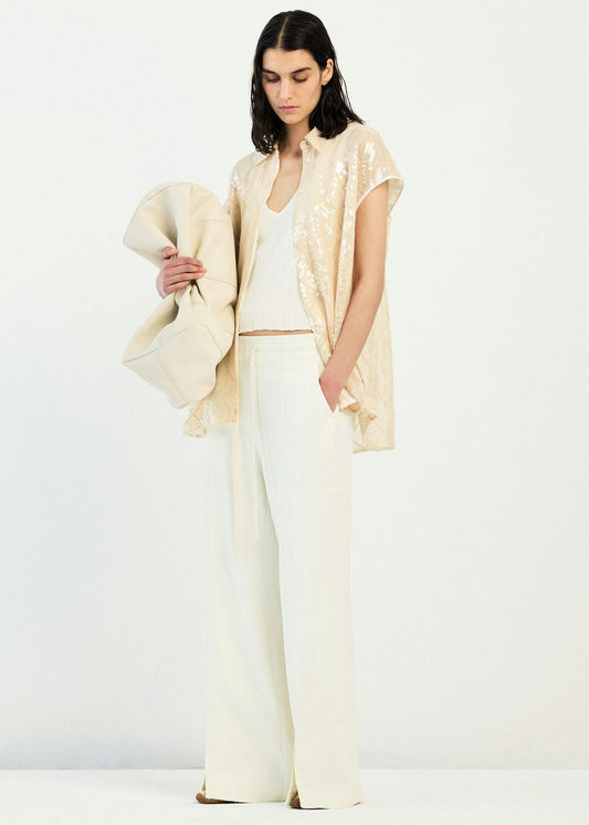 A model stands against a plain white studio background wearing a cream-colored outfit consisting of wide-leg trousers, a white top, a sleeveless cream vest with subtle gold sequins, and holding a matching cream draped wrap.