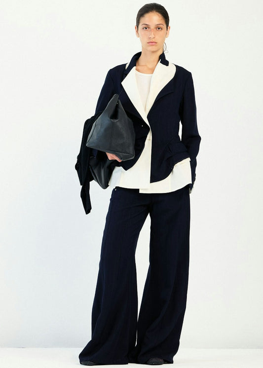 A model stands against a plain white studio background wearing a dark navy wide-leg pants suit with a white shirt underneath, a black blazer draped over one shoulder, and holding a black leather handbag.