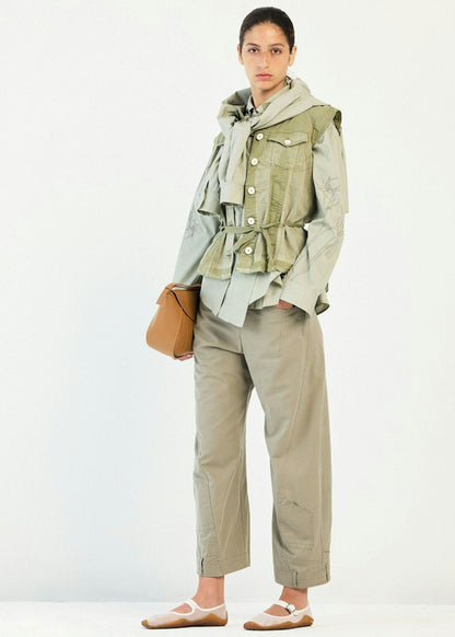 A model stands against a plain white studio background wearing a light olive green ruffled utility jacket over a pale sage green top, paired with matching light olive trousers and white ballet flats, and holding a small tan shoulder bag.