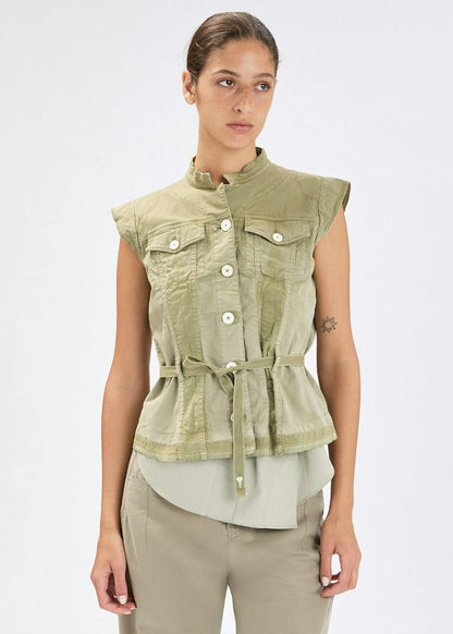 A woman modeling a sleeveless olive green cropped vest with a stand collar, front button closure, chest flap pockets, and a drawstring tie at the waist, paired with light olive pants against a plain light gray studio background.