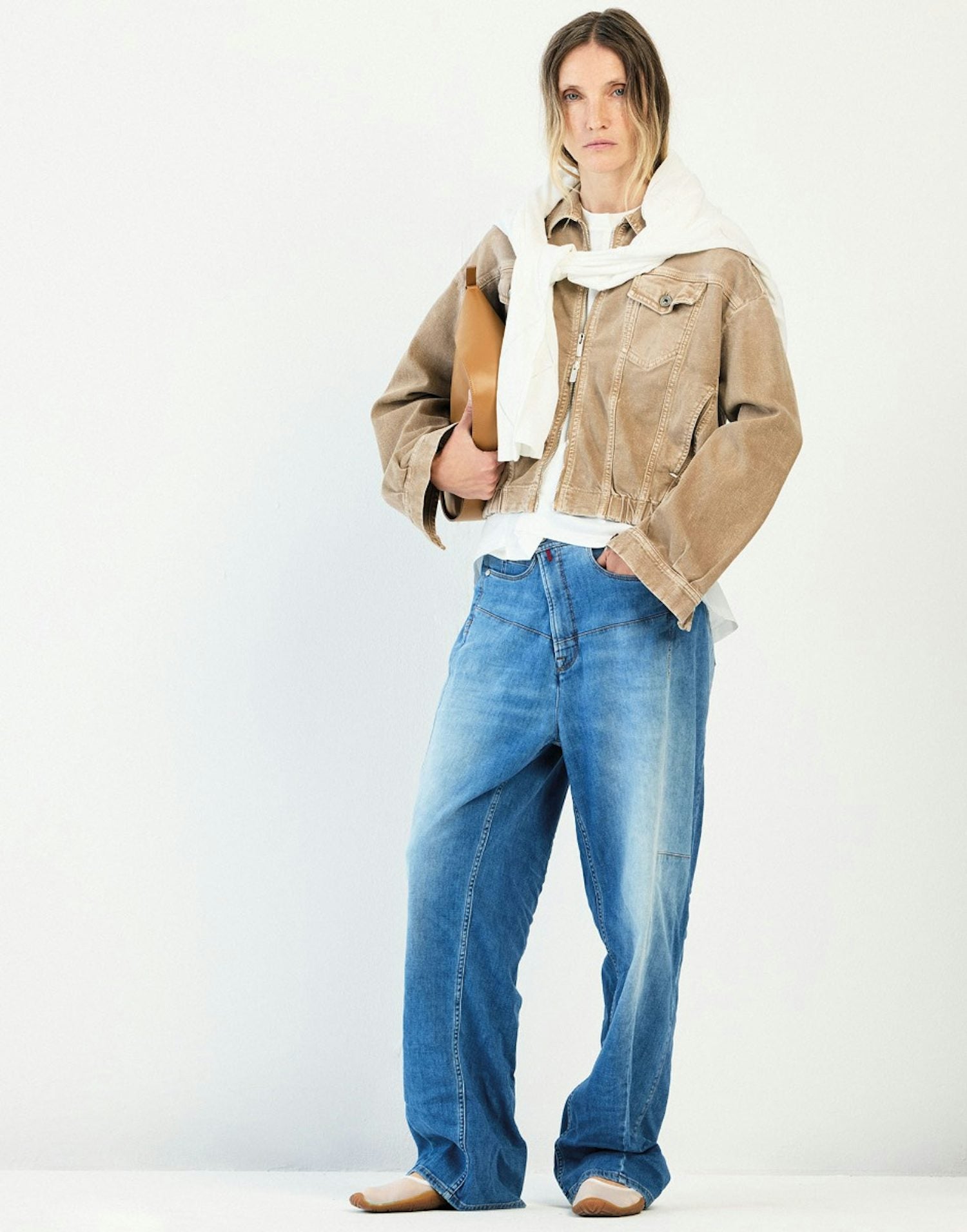 A model stands against a plain white studio background wearing a cropped tan jacket with a white collar, a white top, blue jeans, and beige shoes, while holding a brown leather clutch.