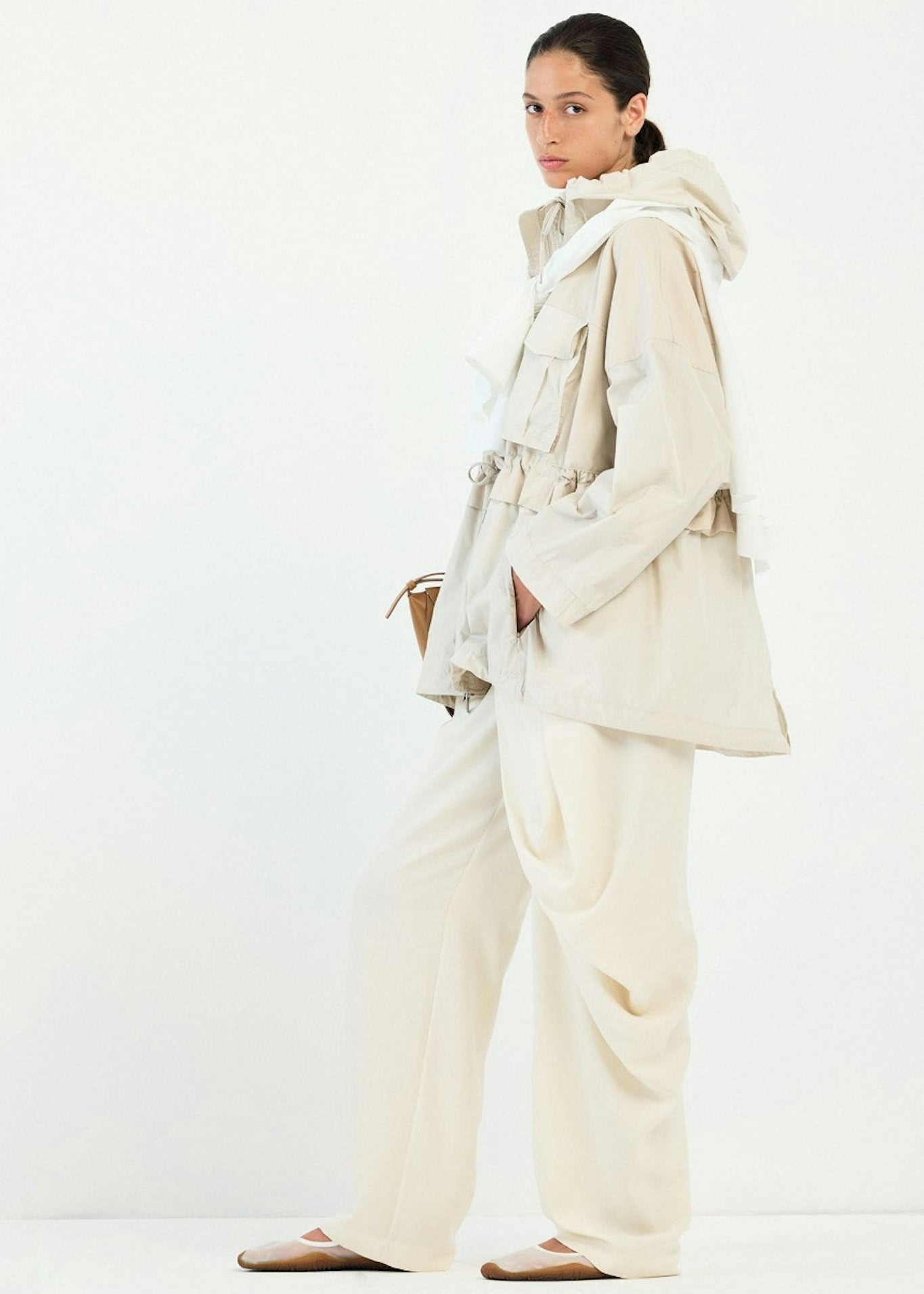 A model wearing a light beige hooded jacket with a tie belt, matching wide-leg trousers, and flat shoes, standing against a plain white studio background.