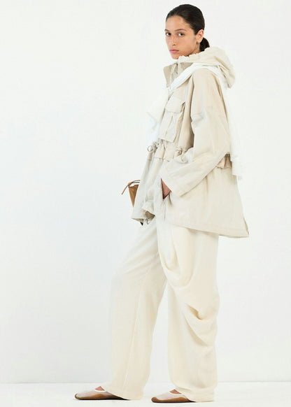 A model wearing a light beige hooded jacket with a tie belt, matching wide-leg trousers, and flat shoes, standing against a plain white studio background.