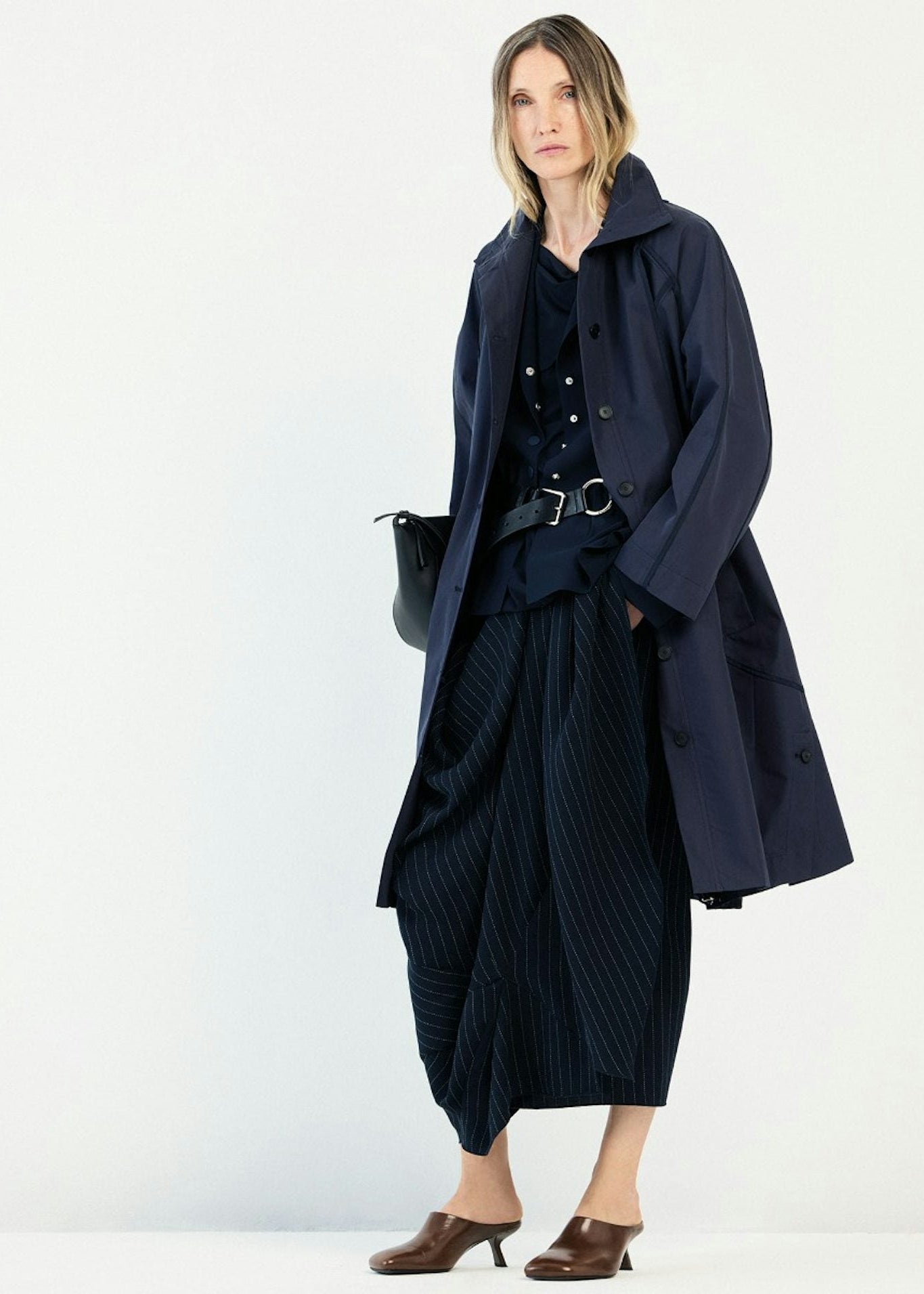 A model stands against a plain white studio background wearing a long navy coat over a matching navy outfit with a pleated skirt, belt, and brown mule heels.