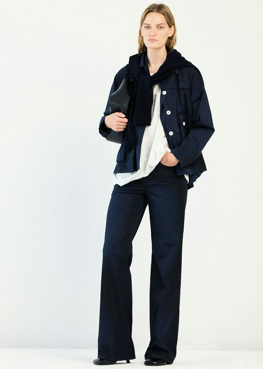 A model stands against a plain white studio background wearing a dark navy jacket with a black collar, a white shirt underneath, dark navy wide-leg trousers, and black shoes.