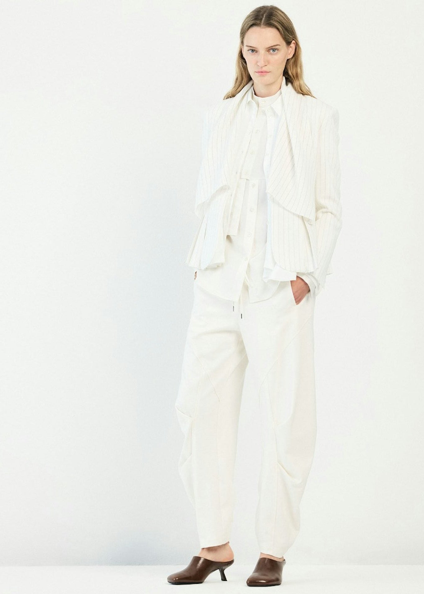 A model wearing a white pinstriped jacket with a high neck and layered ruffle details, paired with matching wide-leg trousers and brown mule heels, set against a plain white studio background.