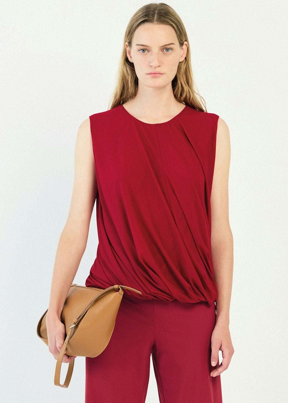 A woman modeling a sleeveless draped top and matching trousers in a deep red color, holding a tan leather crossbody bag against a plain white studio background.