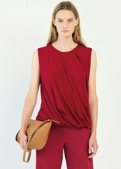 A woman modeling a sleeveless draped top and matching trousers in a deep red color, holding a tan leather crossbody bag against a plain white studio background.