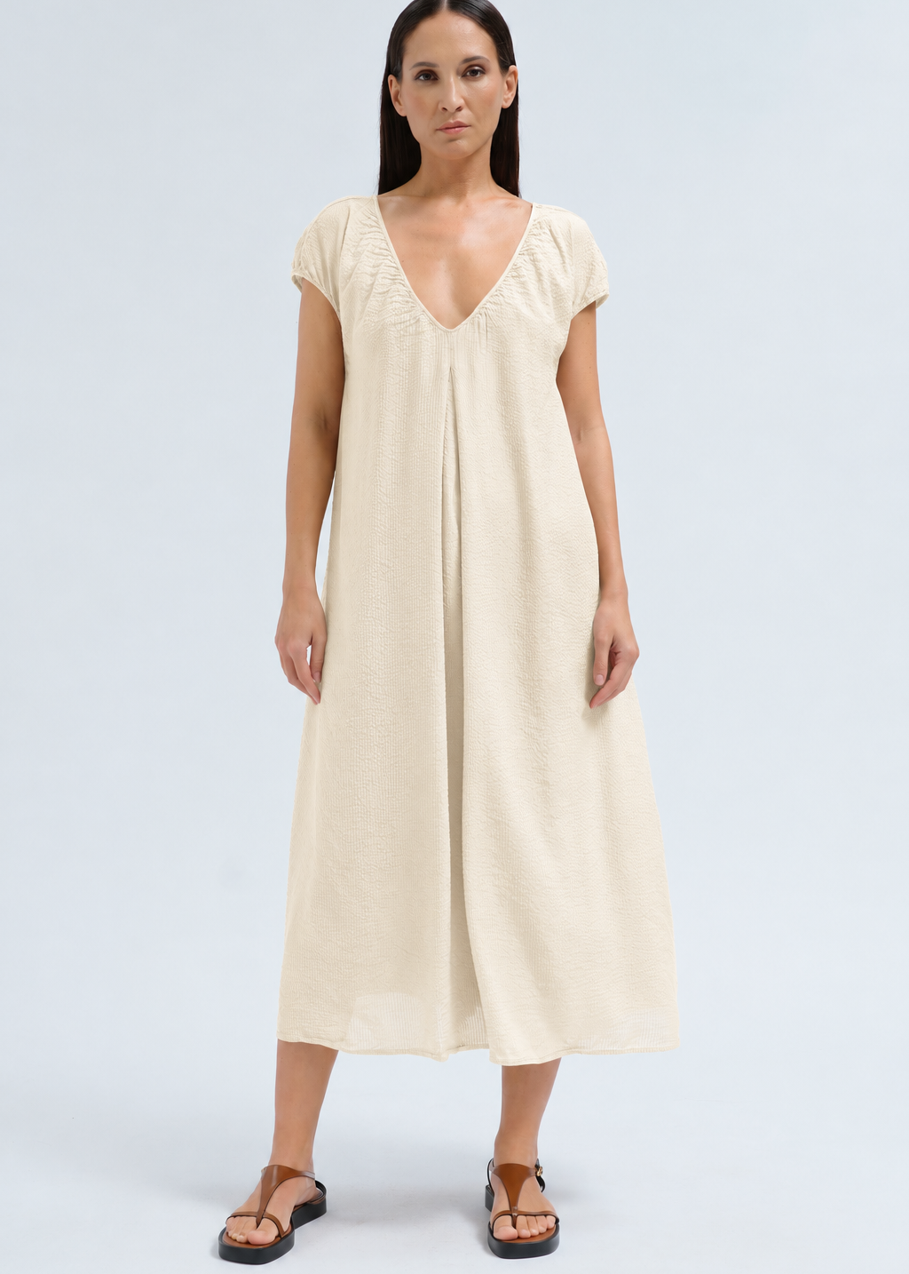 A woman modeling a cream-colored linen midi dress with a V-neckline and cap sleeves, paired with brown leather sandals, against a light gray studio background.