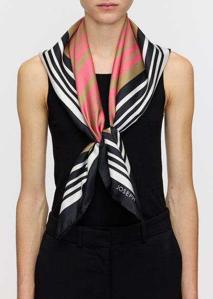 A model wearing a black sleeveless top and black trousers, styled with a Joseph Goldsmith silk scarf tied around the neck. The scarf features a pattern of black, white, pink, and gold stripes.