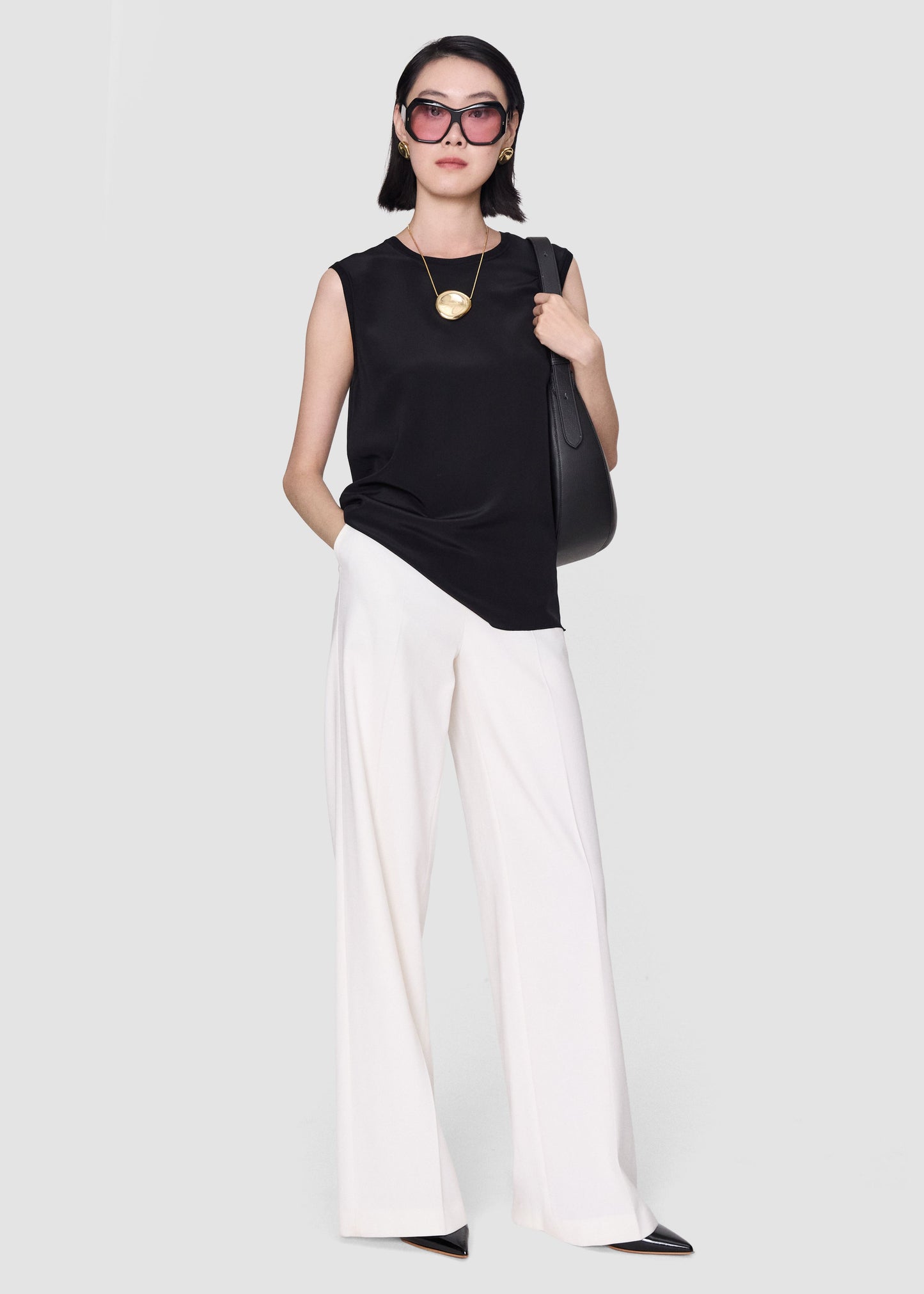 A model wearing a sleeveless black top, white wide-leg pants, black pointed shoes, and black sunglasses, carrying a black shoulder bag against a light gray studio background.