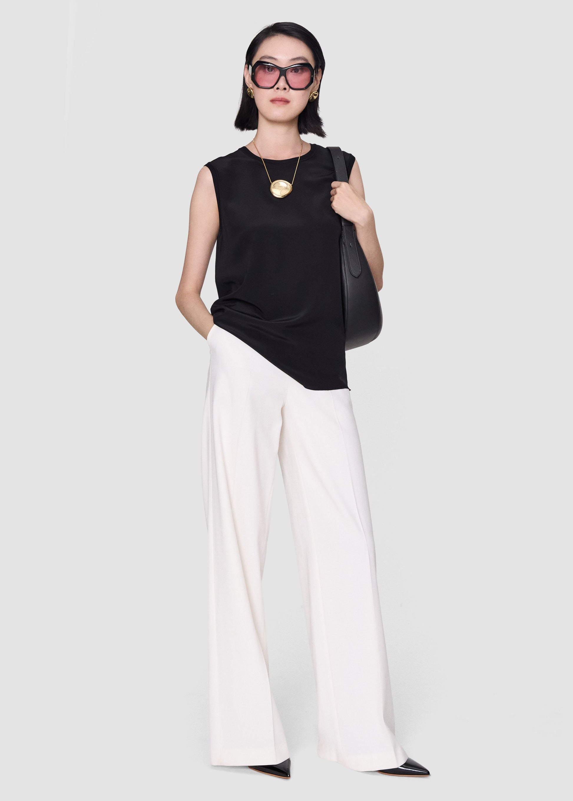 A model wearing a sleeveless black top, white wide-leg pants, black pointed shoes, and black sunglasses, carrying a black shoulder bag against a light gray studio background.
