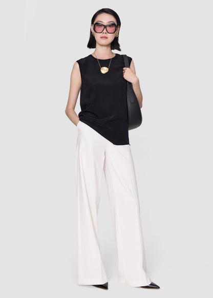 A model wearing a sleeveless black top, white wide-leg pants, black pointed shoes, and black sunglasses, carrying a black shoulder bag against a light gray studio background.