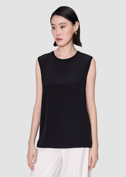 A woman modeling a sleeveless black top with a crew neck, paired with white pants, against a solid light gray background.