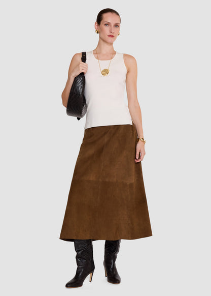 A model stands against a light gray studio background wearing a sleeveless cream top, a brown midi skirt, black knee-high boots, and carrying a black textured shoulder bag.