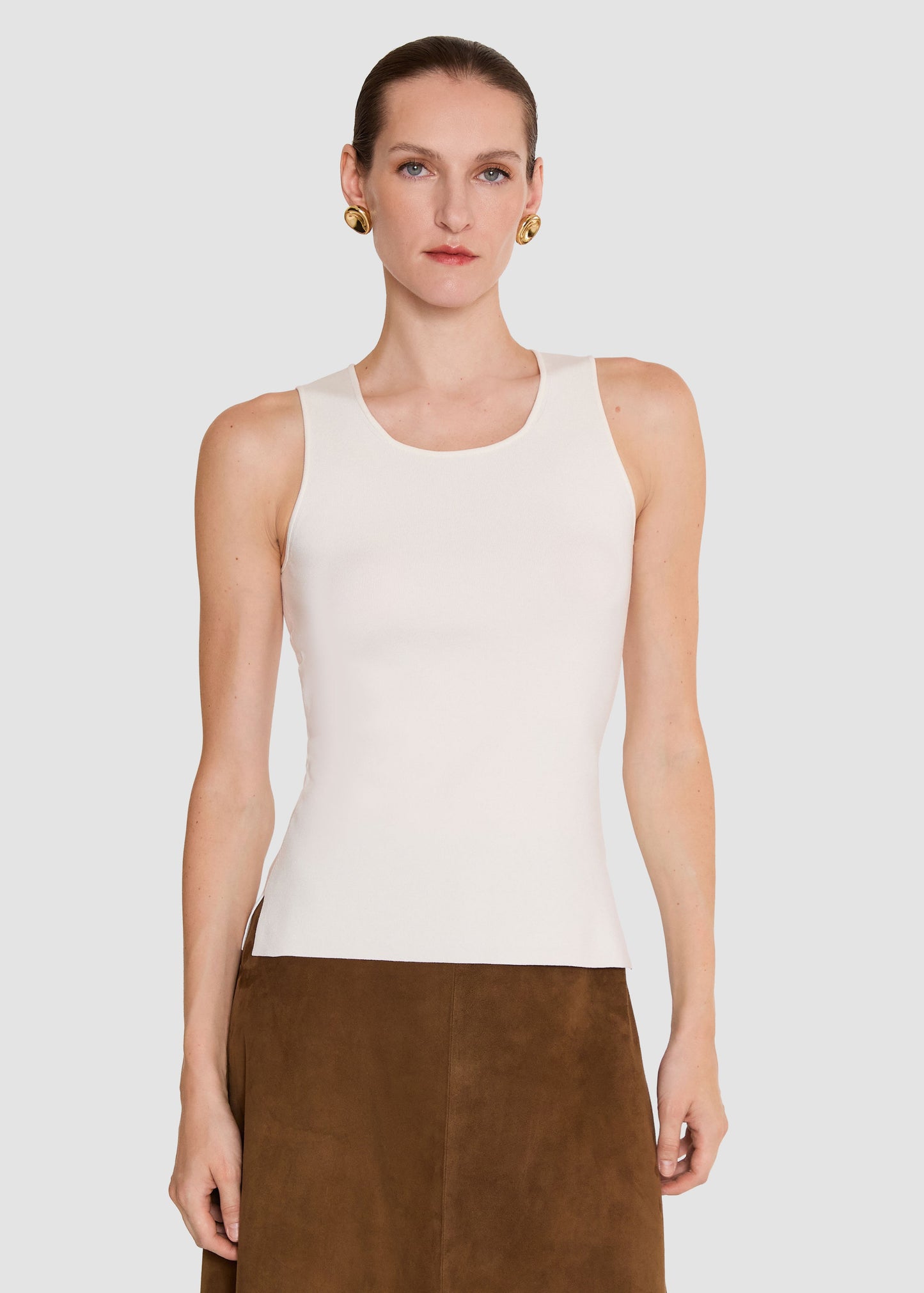 A model wearing a fitted sleeveless knit top in off-white/cream, paired with a brown skirt, against a light gray studio background.