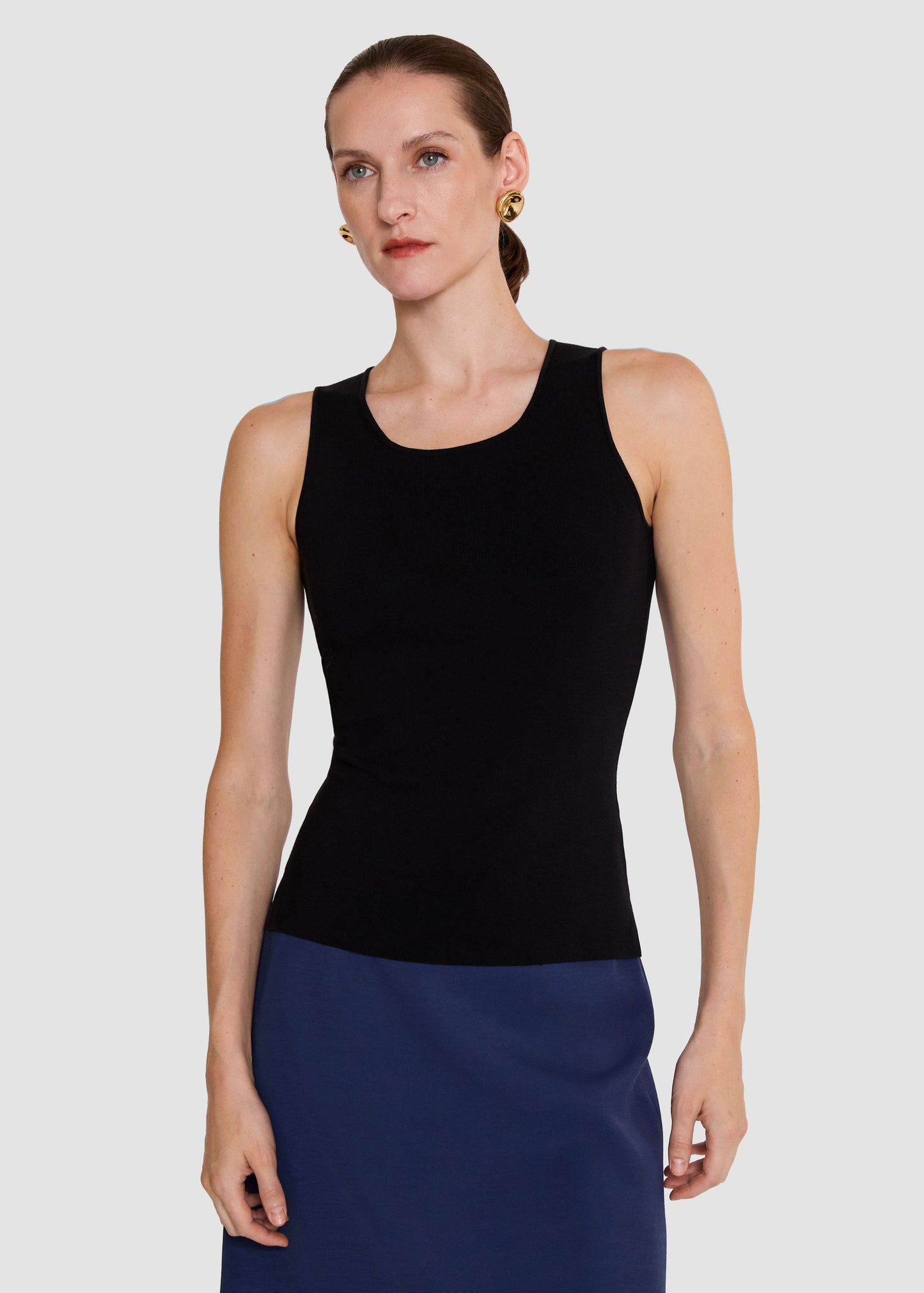 A woman modeling a black sleeveless scoop-neck top paired with a navy blue skirt, photographed against a solid light gray background.