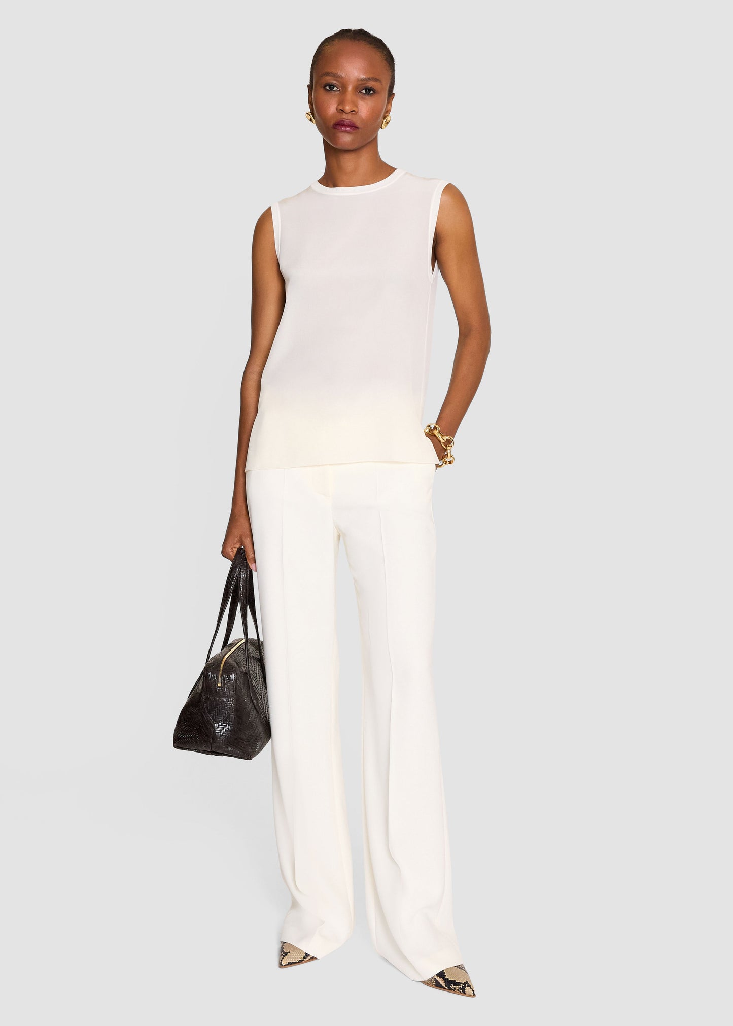 A model stands against a light gray studio background wearing a sleeveless white knit top and matching wide-leg white trousers, holding a dark brown textured handbag.
