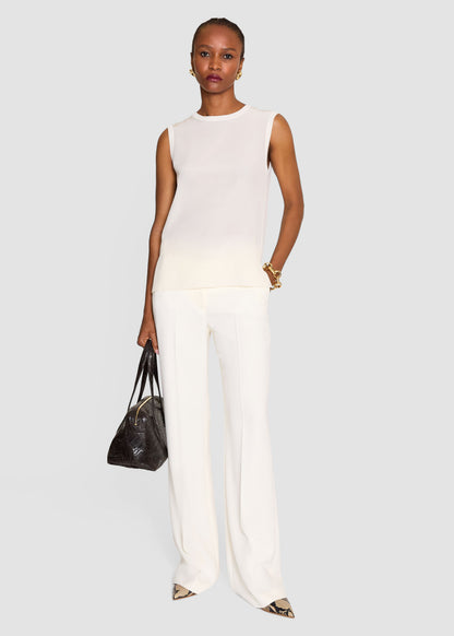 A model stands against a light gray studio background wearing a sleeveless white knit top and matching wide-leg white trousers, holding a dark brown textured handbag.