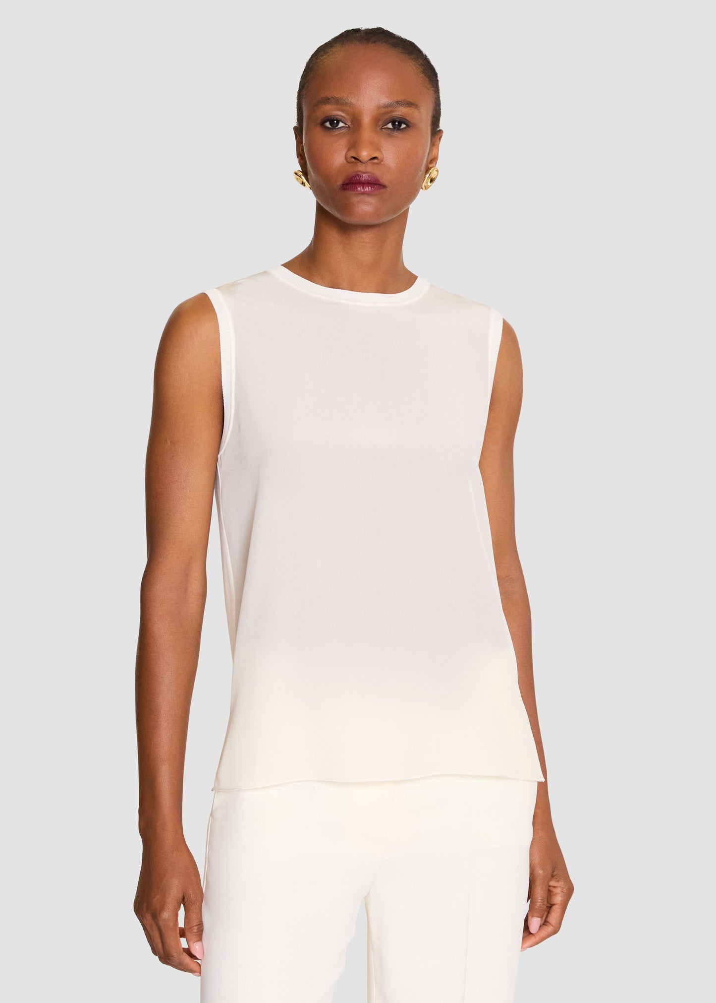 A model wearing a sleeveless, semi-sheer white top and matching white trousers, photographed against a solid light gray background.