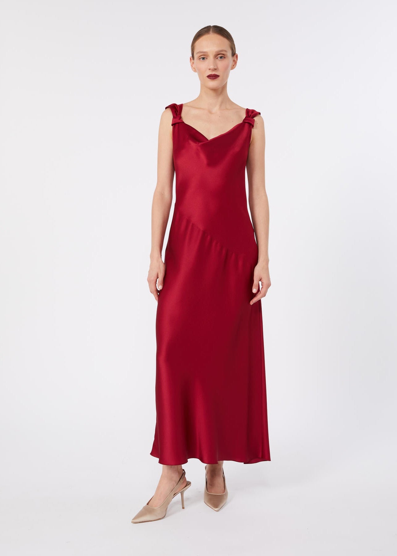A woman modeling a sleeveless, deep red satin maxi dress with shoulder ruffles, paired with beige high heels, against a plain light gray studio background.