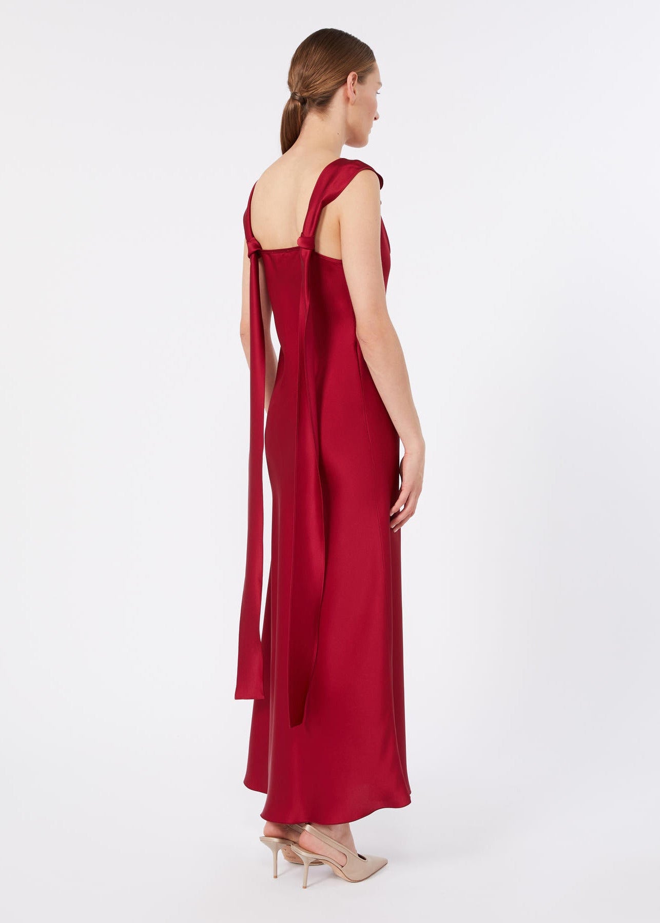 A woman modeling a floor-length red satin gown from the back, featuring a square neckline, wide straps, and a long draped tie detail, set against a plain light gray studio background.