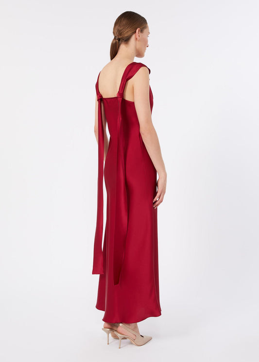 A woman modeling a floor-length red satin gown from the back, featuring a square neckline, wide straps, and a long draped tie detail, set against a plain light gray studio background.