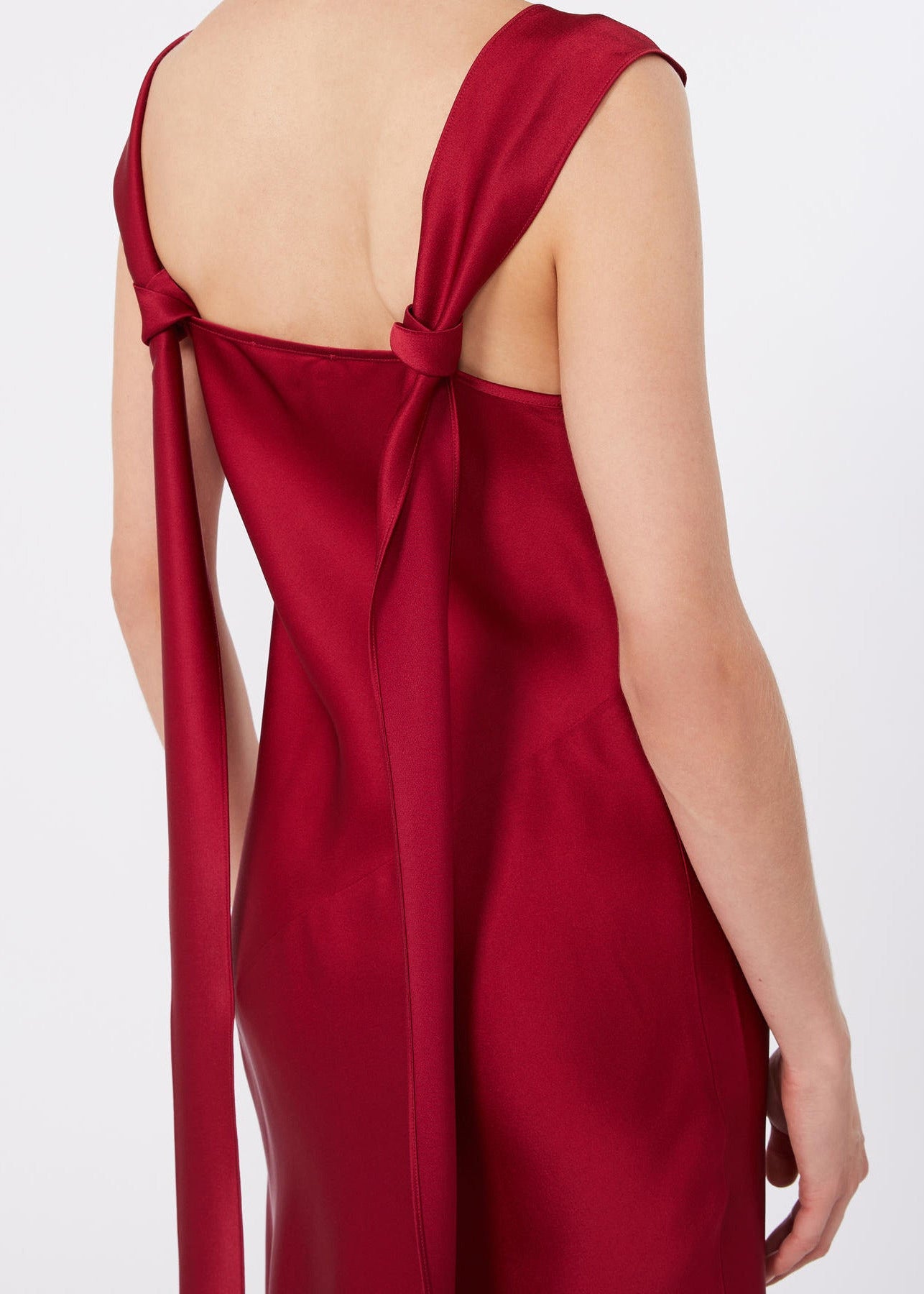 A close-up back view of a woman wearing a satin slip dress in deep red. The dress features a low open back with a bow detail at the top and long tie straps hanging down the sides.