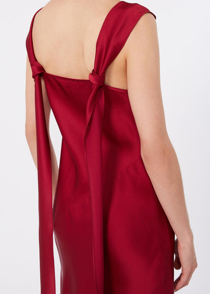A close-up back view of a woman wearing a satin slip dress in deep red. The dress features a low open back with a bow detail at the top and long tie straps hanging down the sides.
