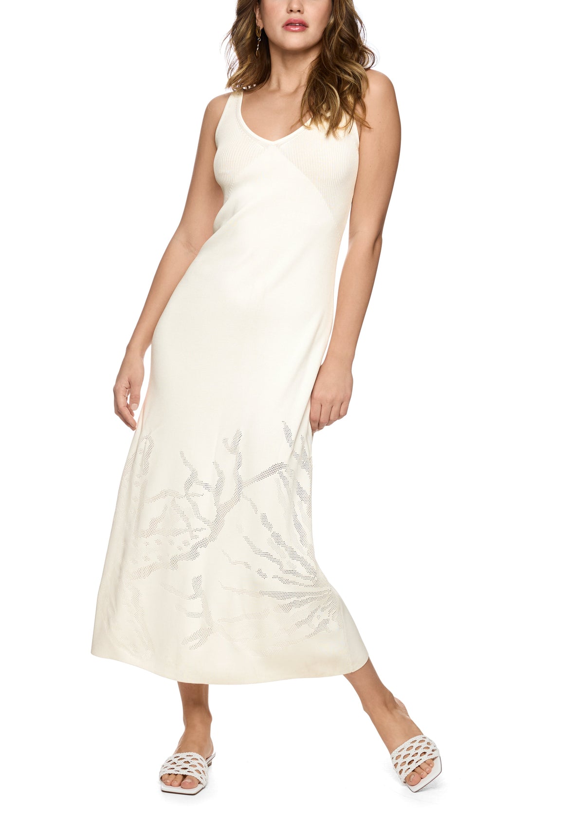 A woman modeling a sleeveless cream maxi dress with a V-neckline and a tonal branch or coral-like pattern near the lower skirt, paired with white strappy sandals against a plain white background.
