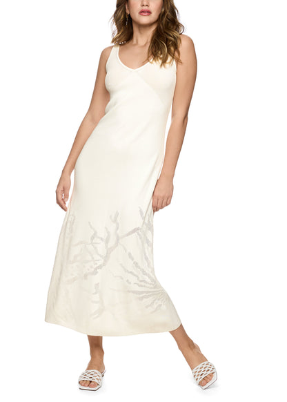 A woman modeling a sleeveless cream maxi dress with a V-neckline and a tonal branch or coral-like pattern near the lower skirt, paired with white strappy sandals against a plain white background.