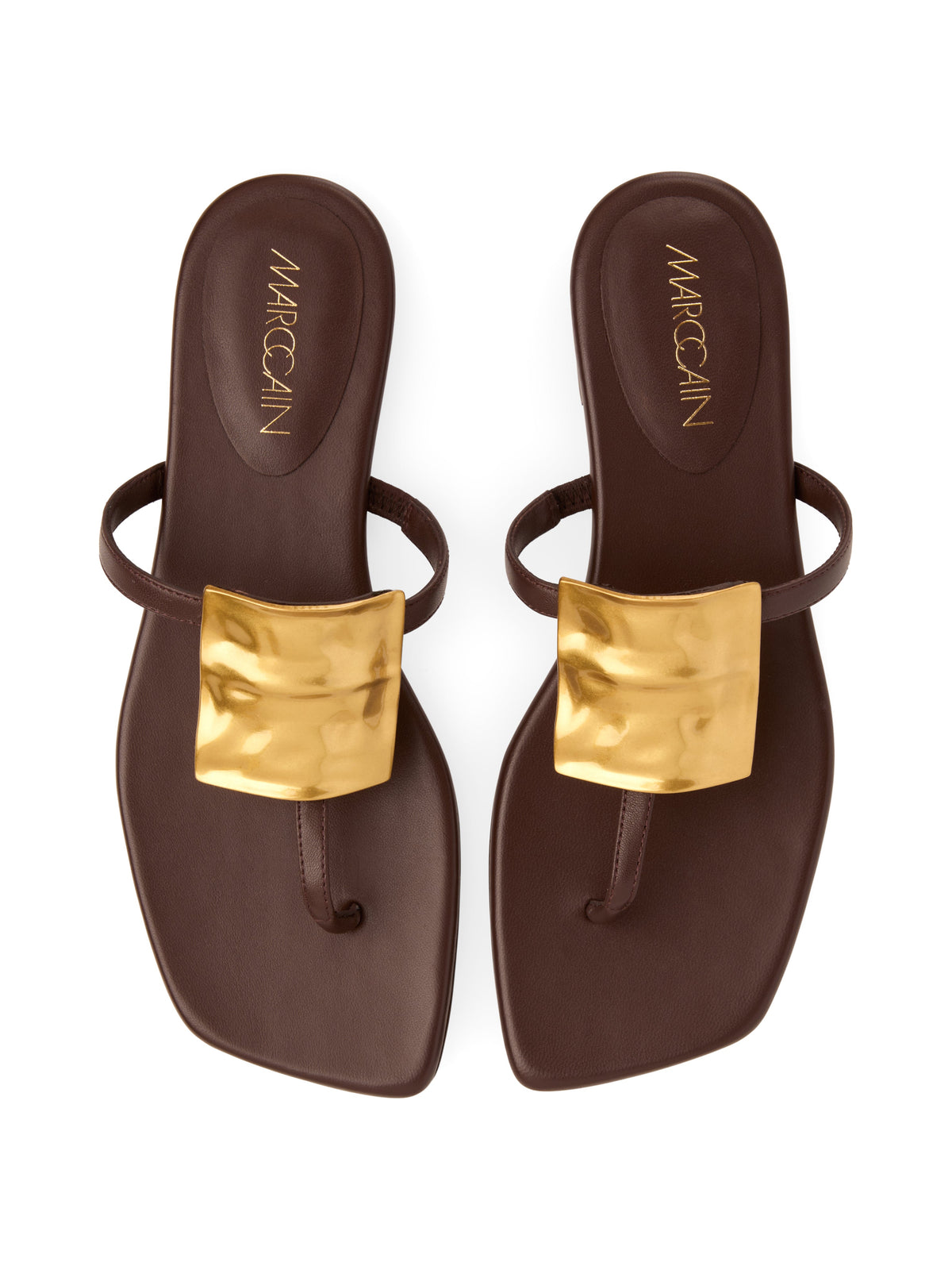 A pair of dark brown flat thong sandals with metallic gold square buckles on the straps, shown from a top-down view on a white background. The brand name 'MARCCAIN' is printed in gold on the heel of each sandal.