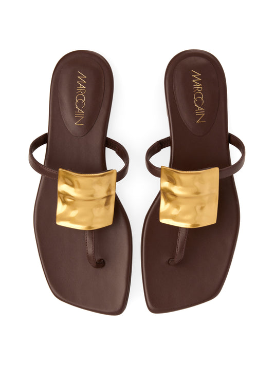 A pair of dark brown flat thong sandals with metallic gold square buckles on the straps, shown from a top-down view on a white background. The brand name 'MARCCAIN' is printed in gold on the heel of each sandal.