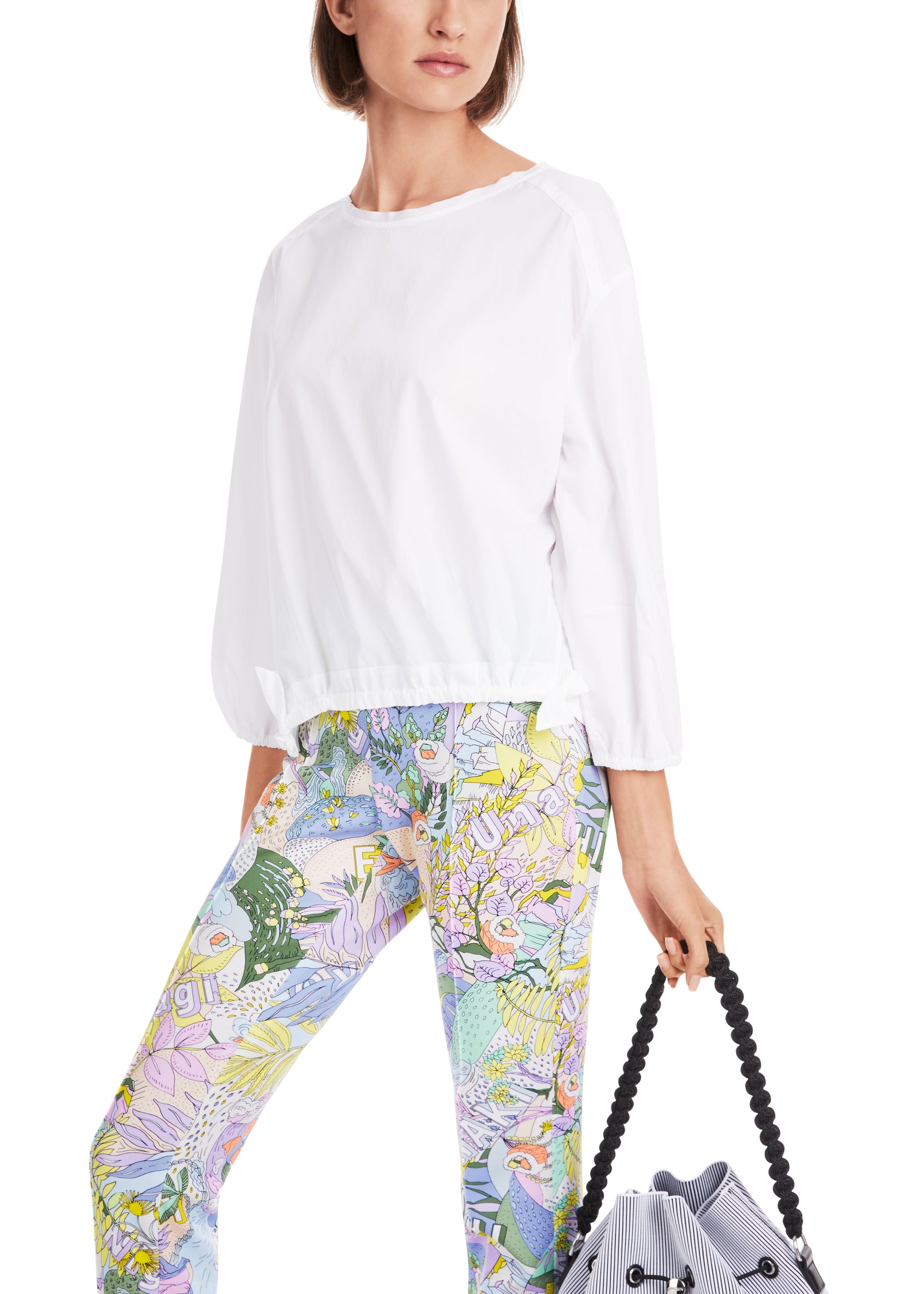 Woman wearing a white top and colorful floral pants holding a gray bag.
