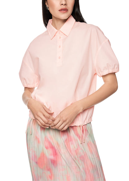 A woman wearing a light pink short-sleeve button-up shirt with a collar and gathered elastic hem, paired with a pastel patterned skirt, against a plain white background.