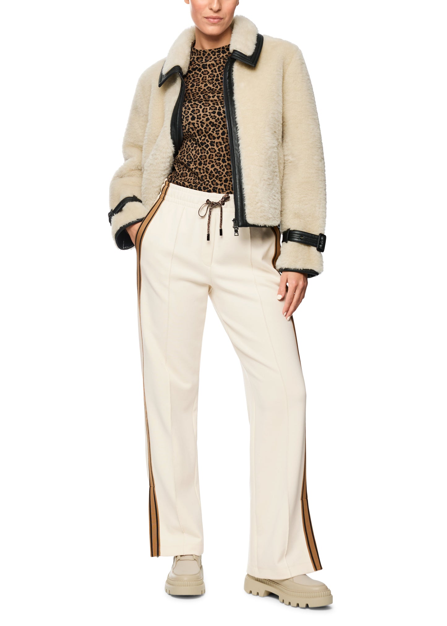 Person wearing a beige shearling jacket, leopard print top, and white pants on a white background