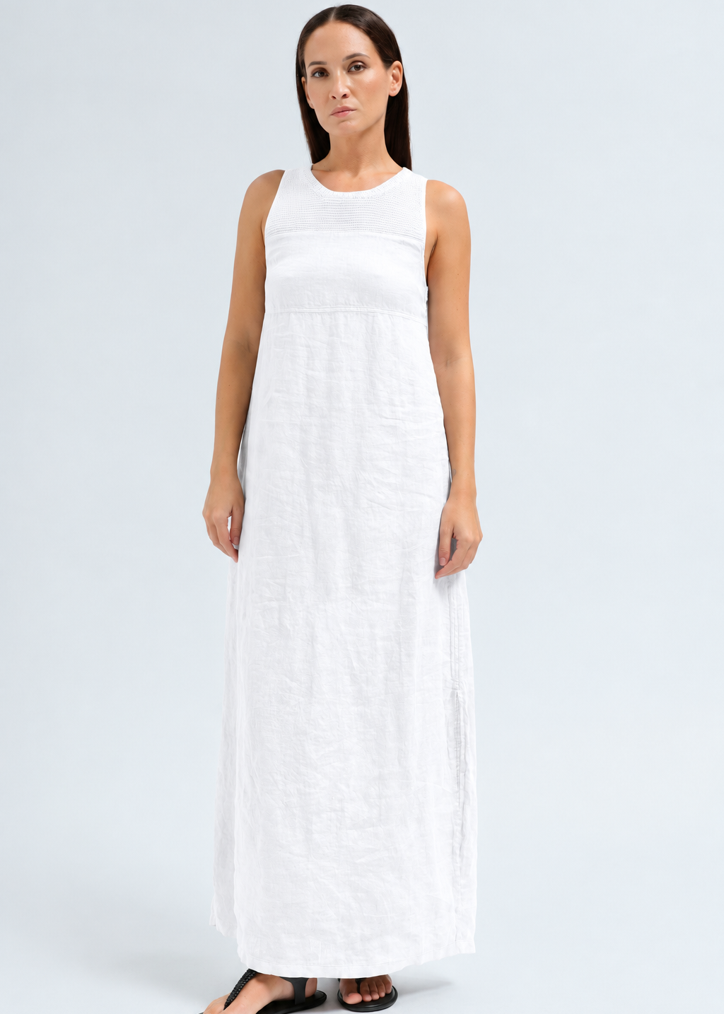 A woman modeling a sleeveless white maxi dress with a textured linen-like fabric, standing against a light gray studio background.