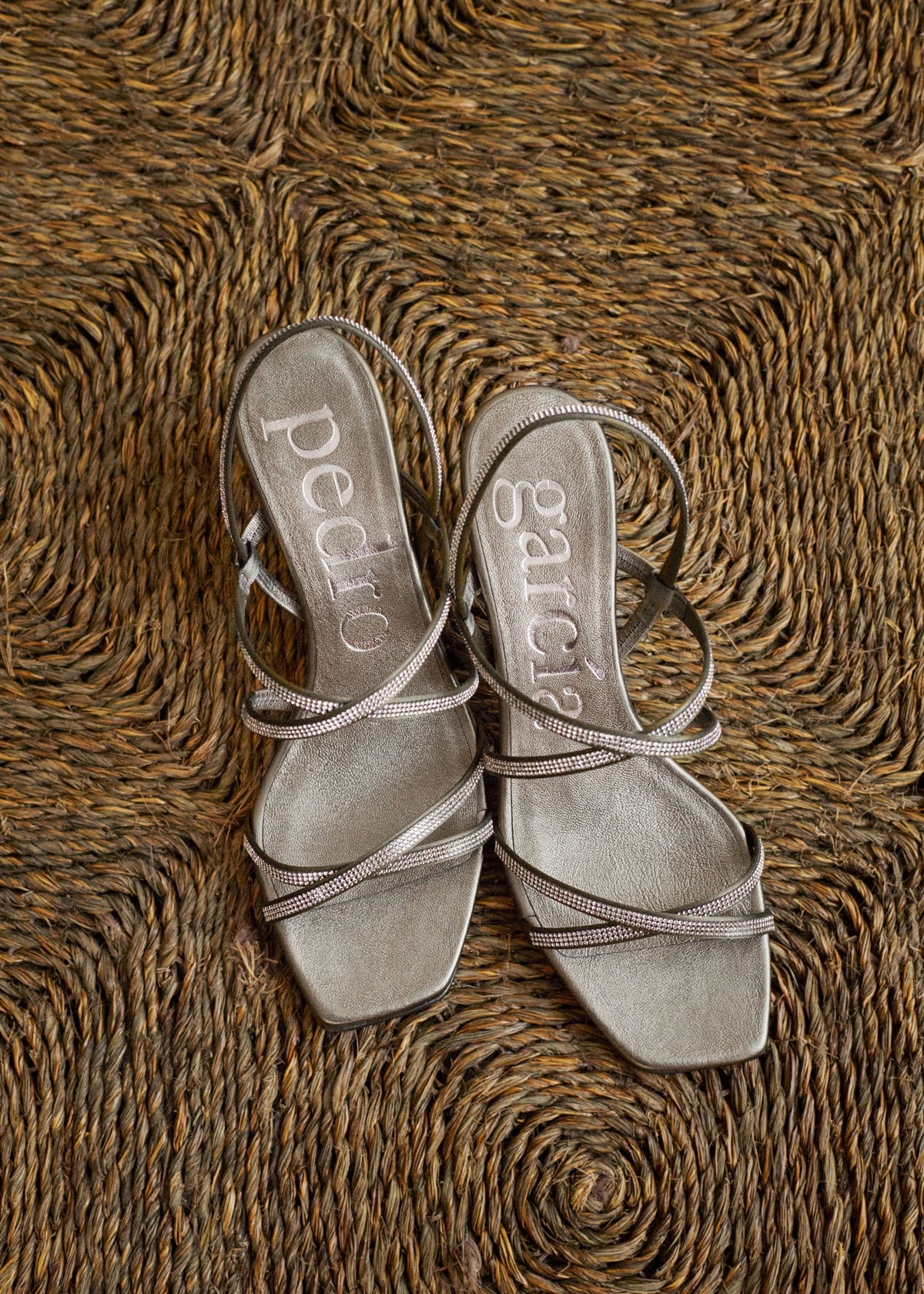 A pair of metallic silver strappy sandals with square toes and low block heels, laid flat on a textured brown woven rug. The brand names 'pedro' and 'sculpe' are printed on the insoles.