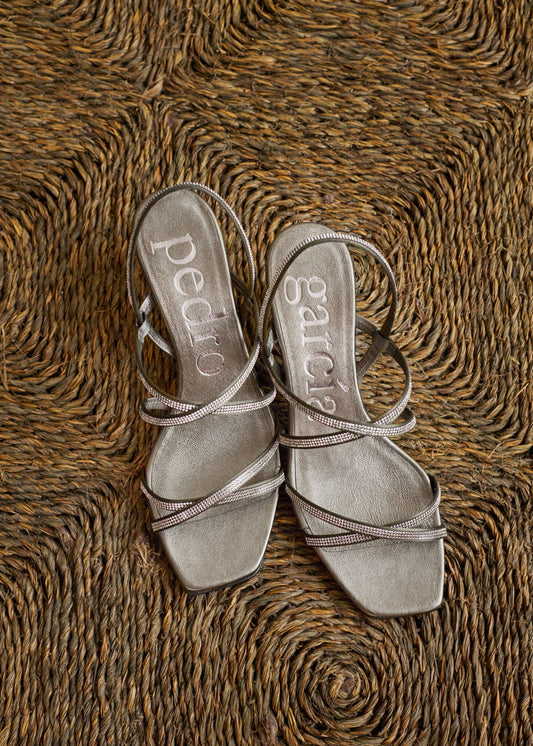 A pair of metallic silver strappy sandals with square toes and low block heels, laid flat on a textured brown woven rug. The brand names 'pedro' and 'sculpe' are printed on the insoles.
