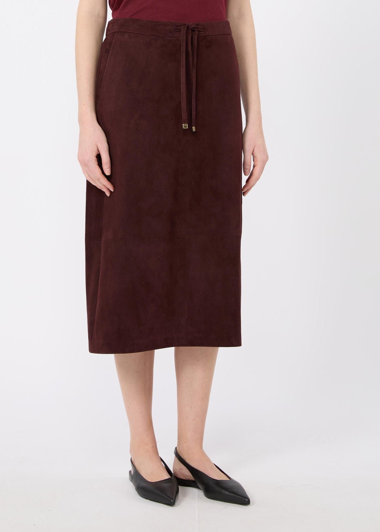 A model wearing a dark brown suede midi skirt with a drawstring waist, paired with black pointed flats, against a plain light gray background.
