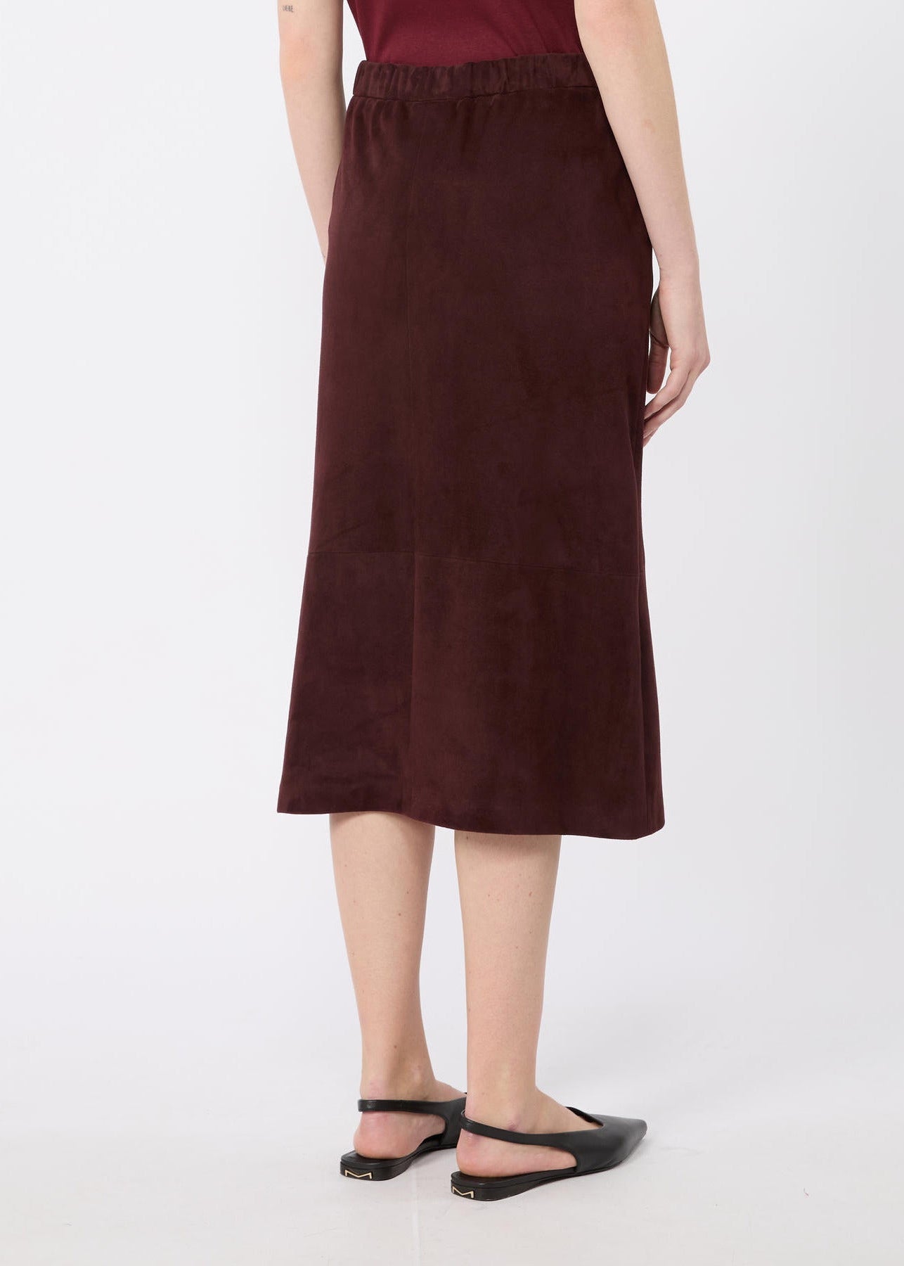 A woman wearing a dark brown midi skirt with an elastic waistband, paired with black flat sandals, shown from the back against a light gray studio background.