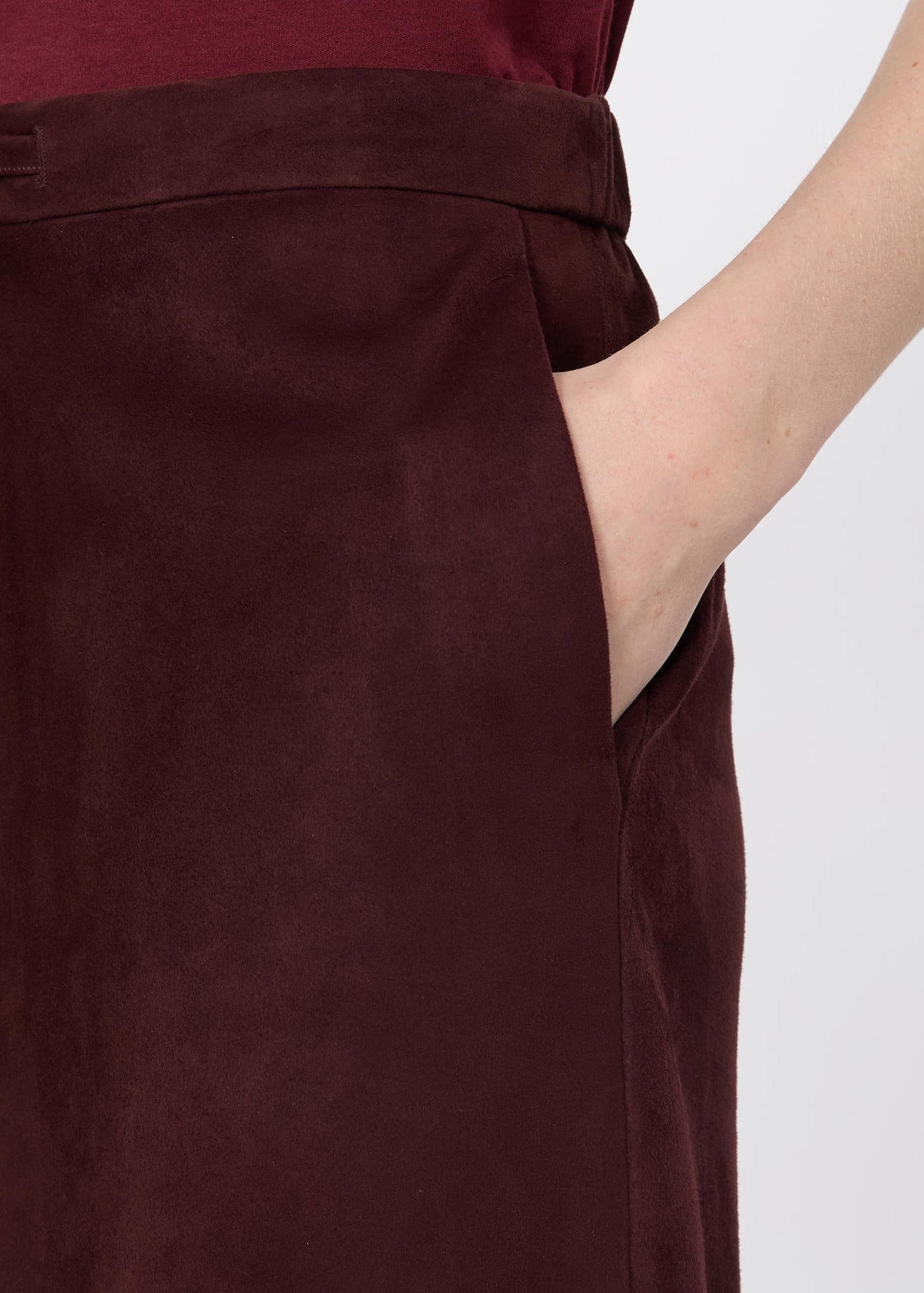 Close-up of a person wearing a dark burgundy skirt or long pants with a side pocket, showing a hand in the pocket against a light gray background.