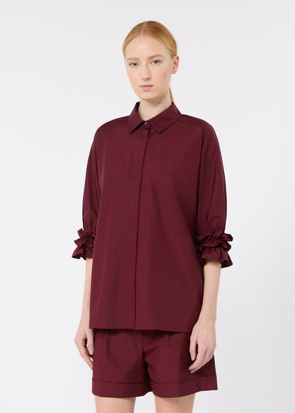 A model wearing a matching burgundy two-piece set consisting of a long-sleeve button-up shirt with ruffled cuffs and matching shorts, shot against a light gray studio background.