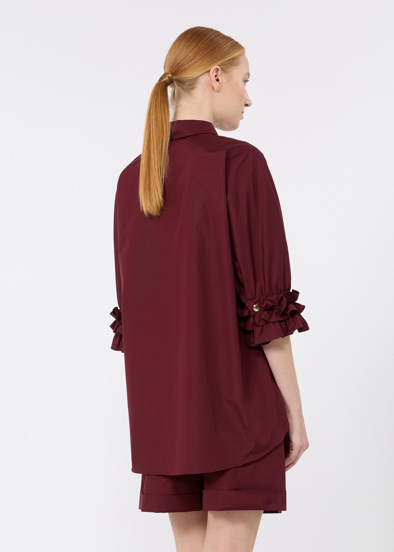 A woman with red hair in a ponytail is shown from the back wearing a burgundy short-sleeve shirt with ruffled cuffs and matching shorts against a light gray studio background.