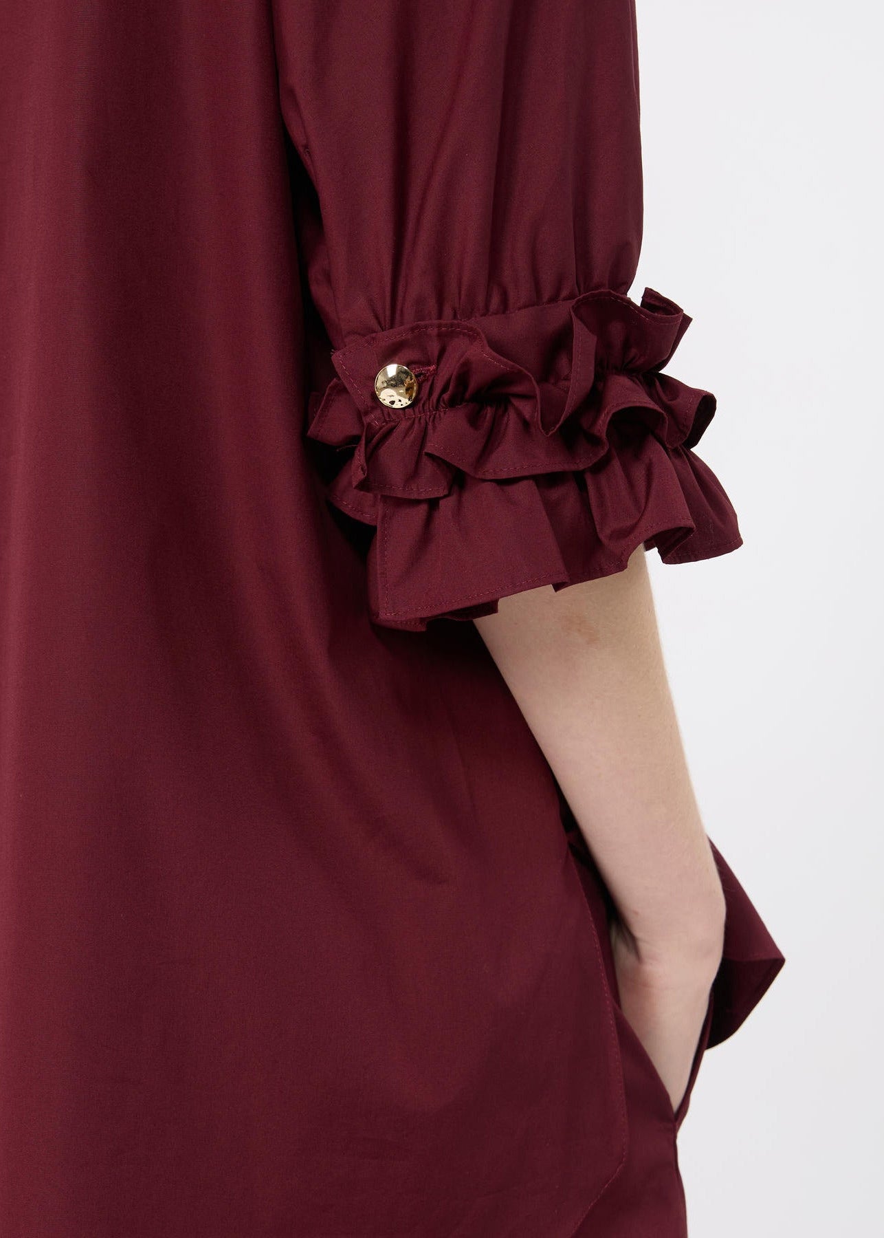 Close-up of a burgundy dress sleeve with a gathered ruffled cuff and a small gold button detail, shown on a model against a light gray background.
