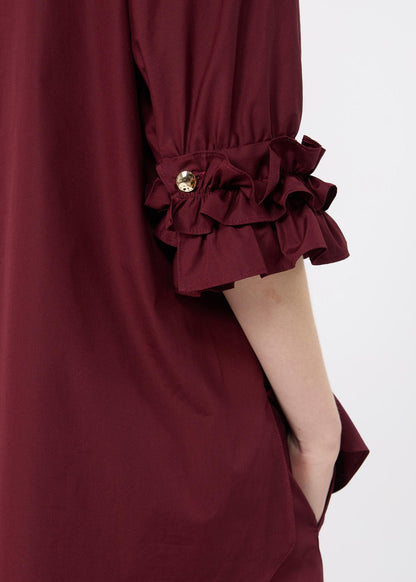 Close-up of a burgundy dress sleeve with a gathered ruffled cuff and a small gold button detail, shown on a model against a light gray background.